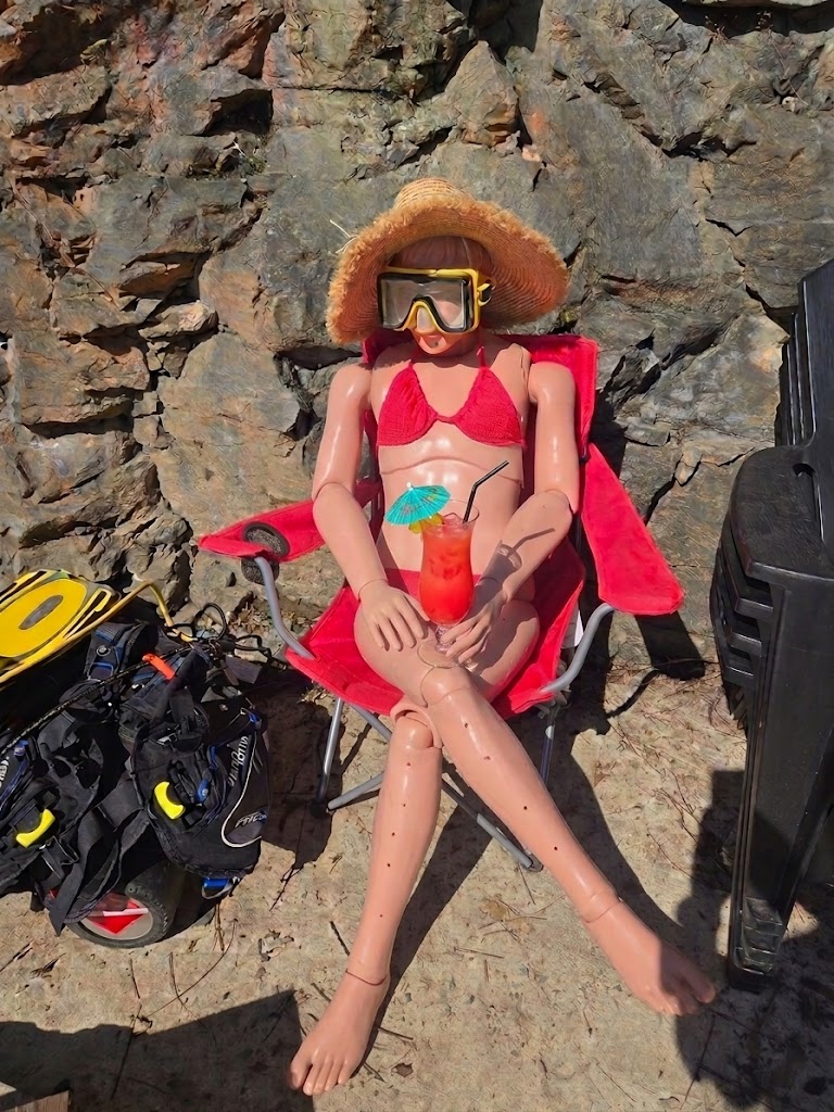 Peggy Sue, a rescue training manikin in a sun hat, dive mask, and bikini, holding a tropical cocktail in a camp chair at the quarry.
